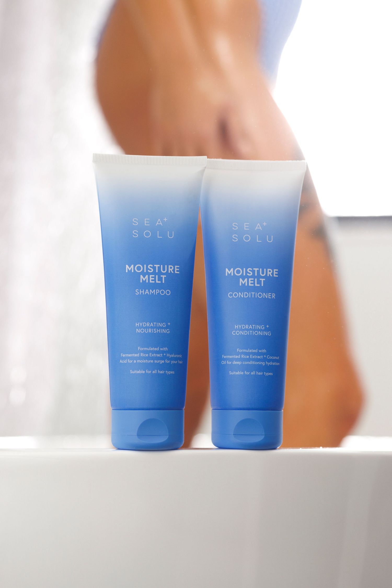 Blue Shampoo & Conditioner tubes arranged minimally on white bathroom surface with someone blurred showering in the background.