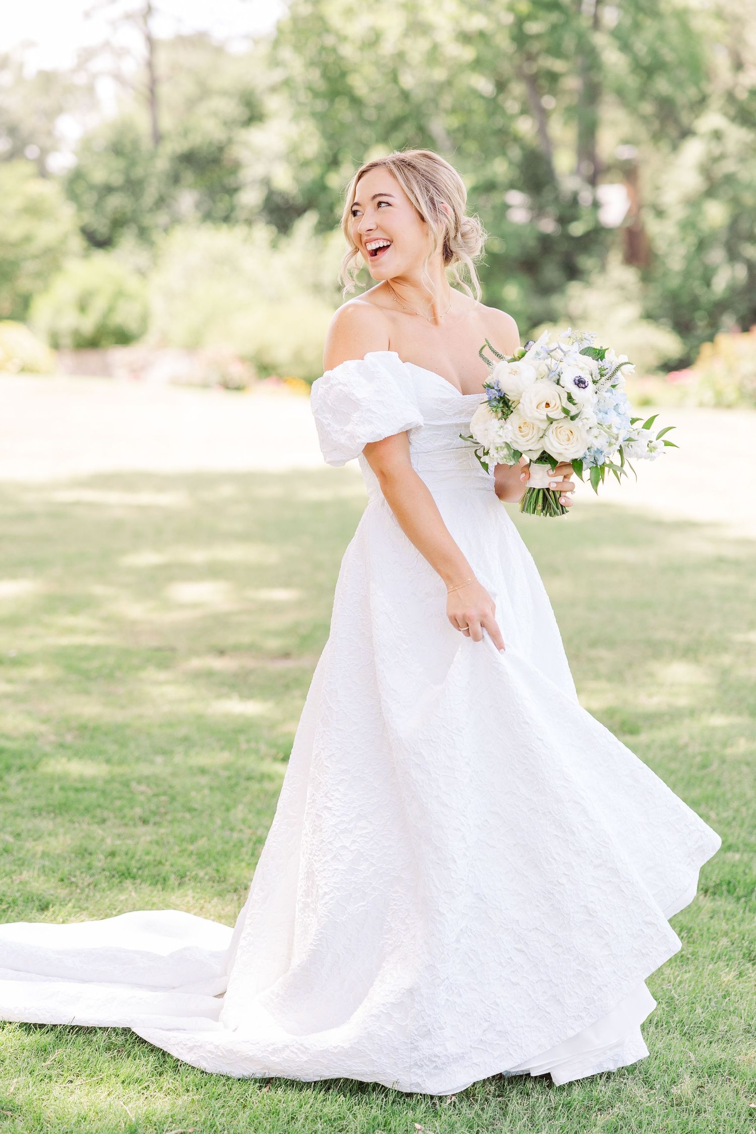Bride in off-shoulder white wedding dress holds white and green bouquet while standing on grass in outdoor garden setting.