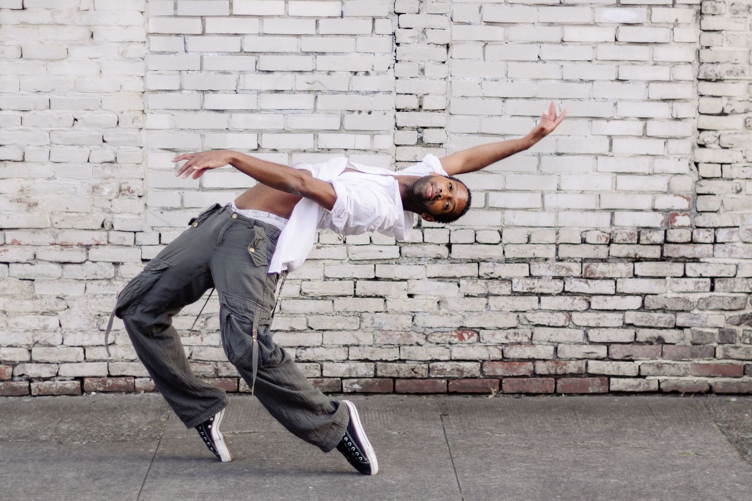 Urban dancer performing a dynamic backbend move against a white brick wall while wearing casual street attire.