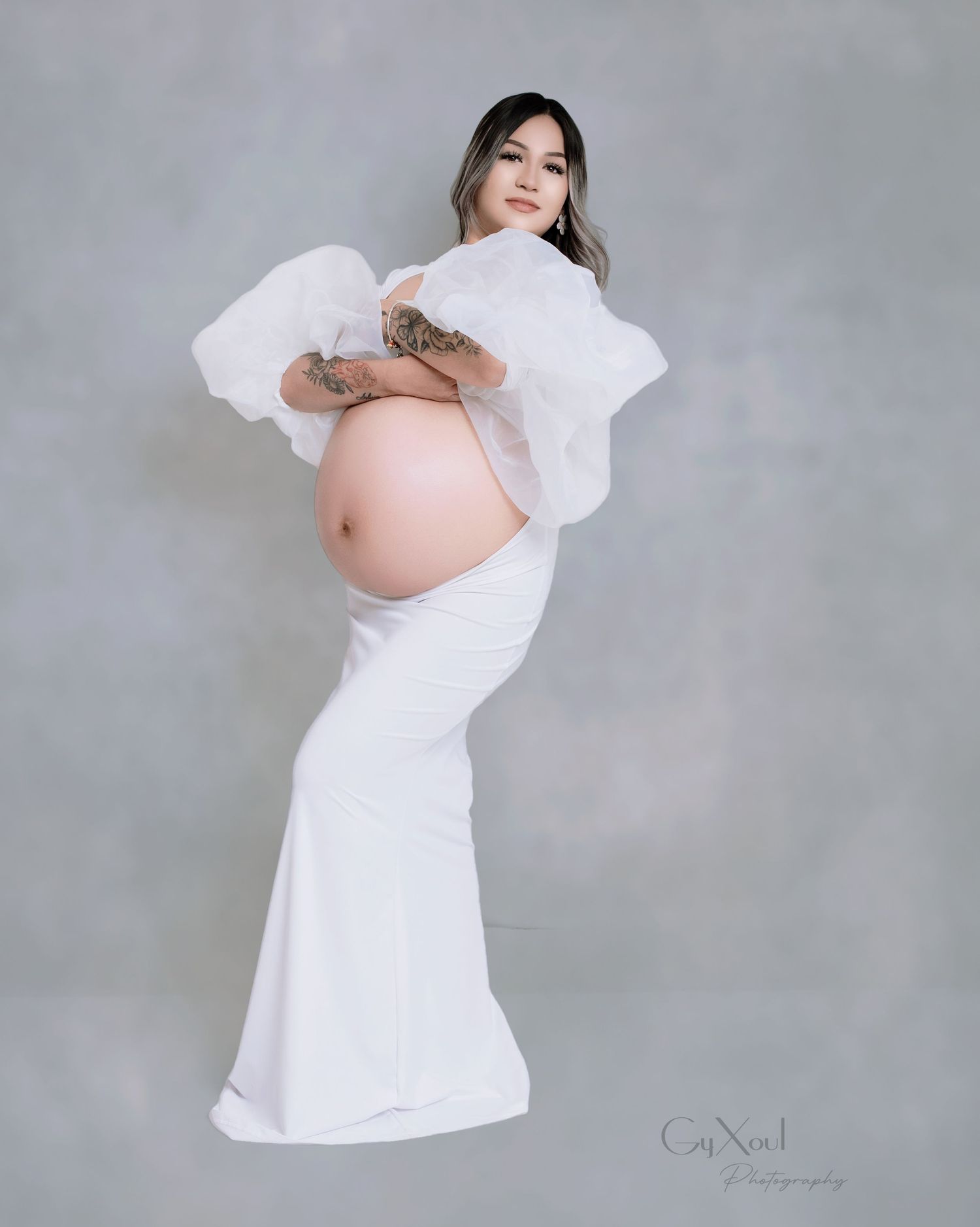 Artistic maternity portrait in flowing white gown against grey backdrop.