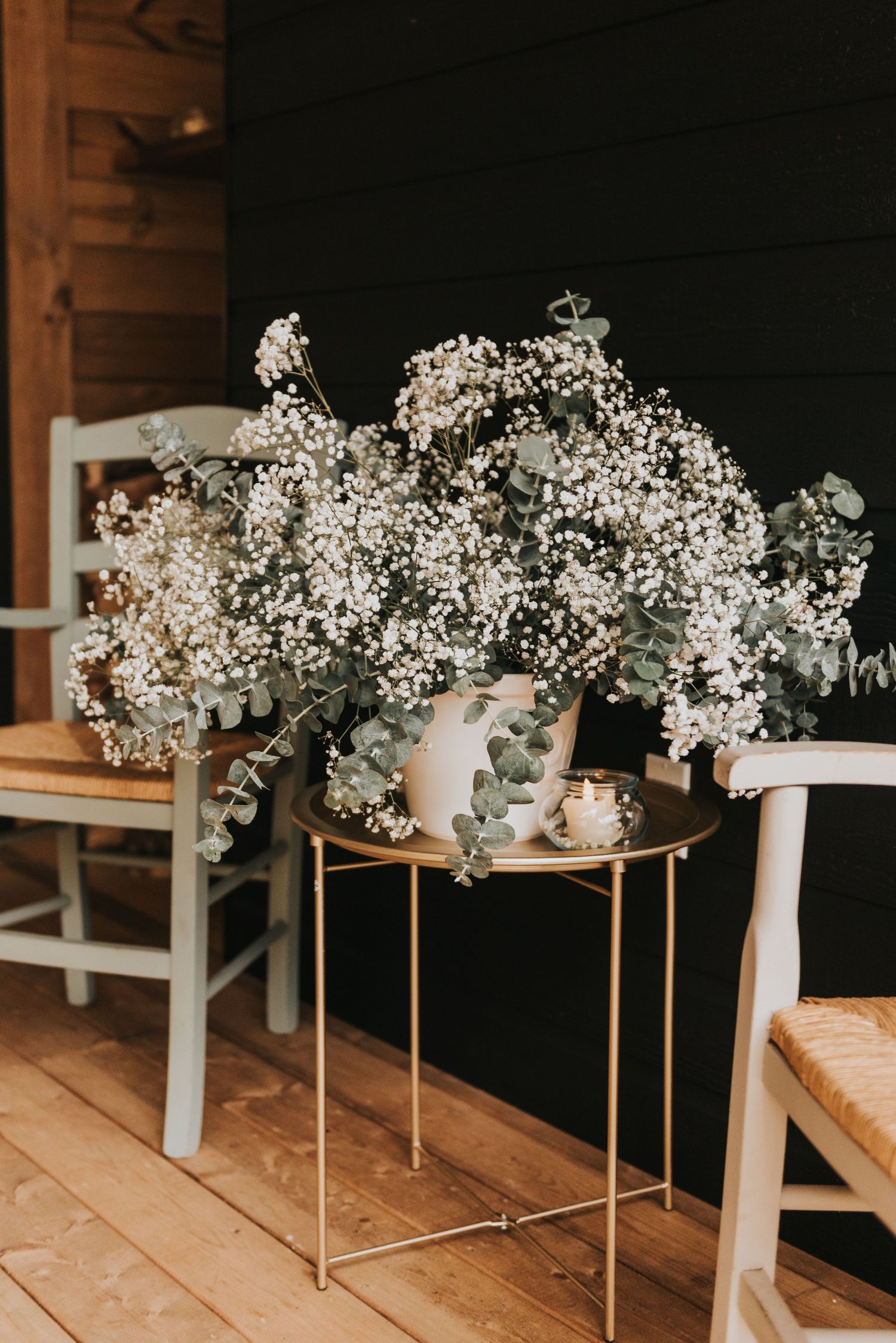 Delicate baby's breath and eucalyptus floral arrangement in white vase on gold side table against wooden wall.
