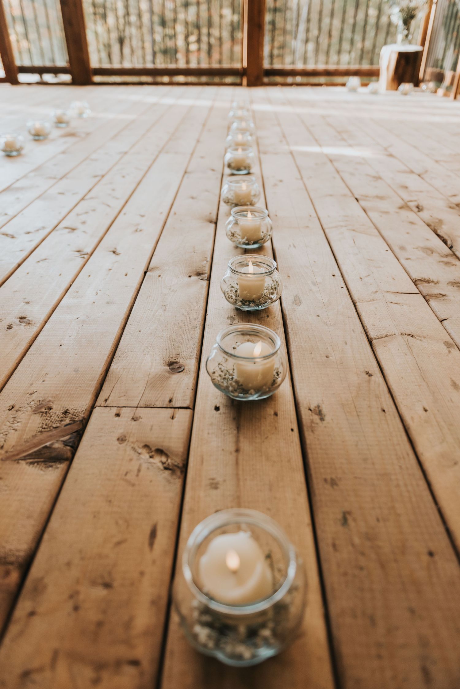 Small glass candle holders with white candles line up along a wooden deck plank in a rustic outdoor setting.