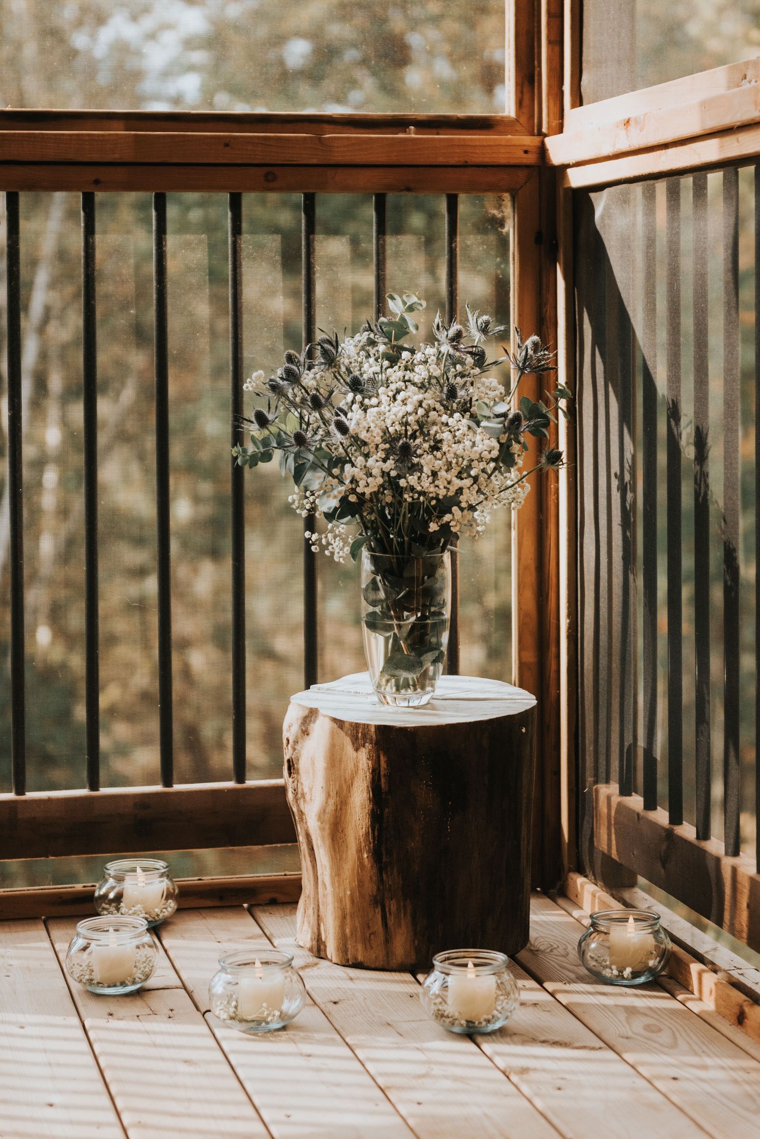 White floral arrangement in glass vase sits atop rustic wood stump on cozy screened porch with scattered tea lights.