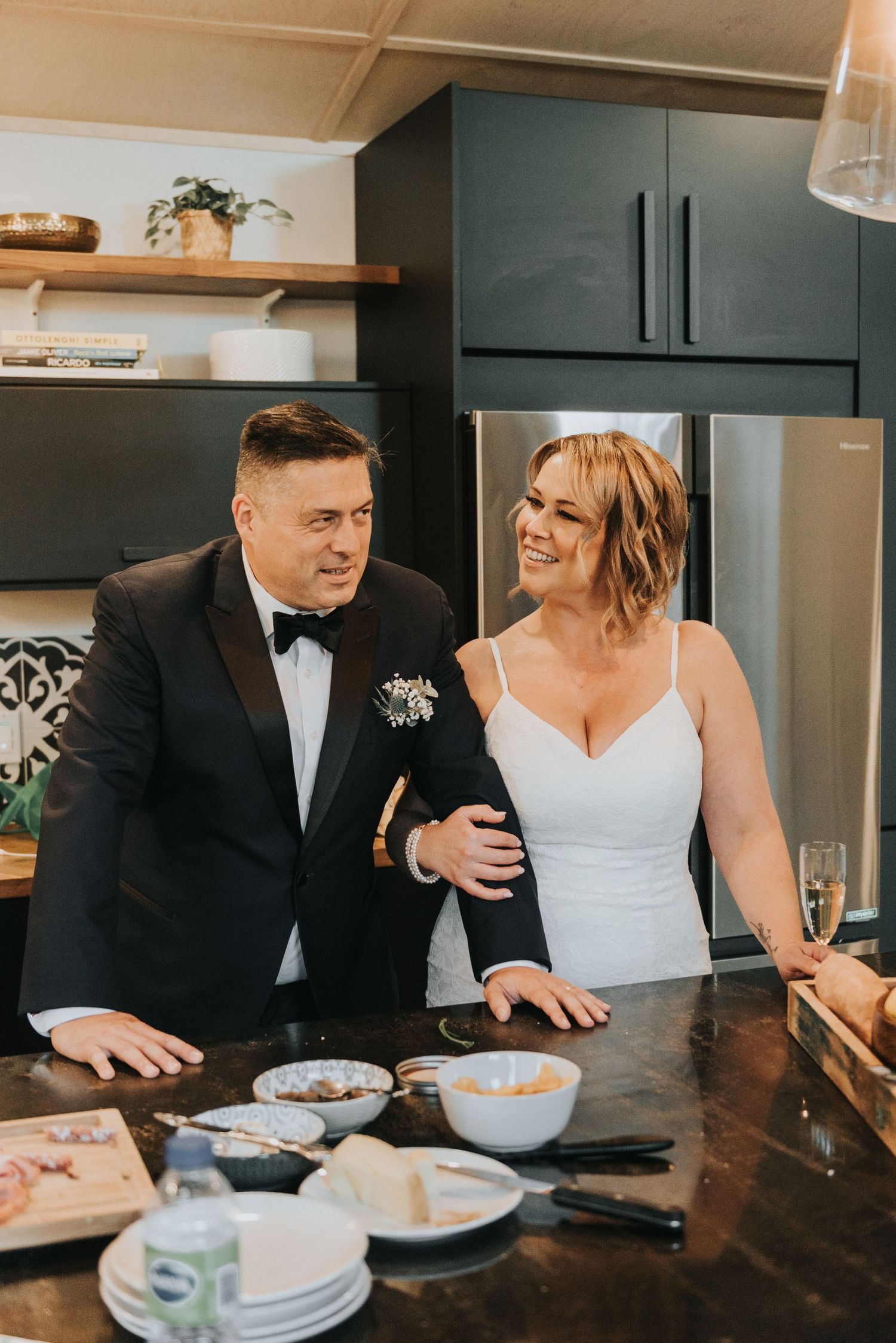 A couple dressed in formal attire sharing a joyful moment in a modern kitchen during what appears to be their wedding day.