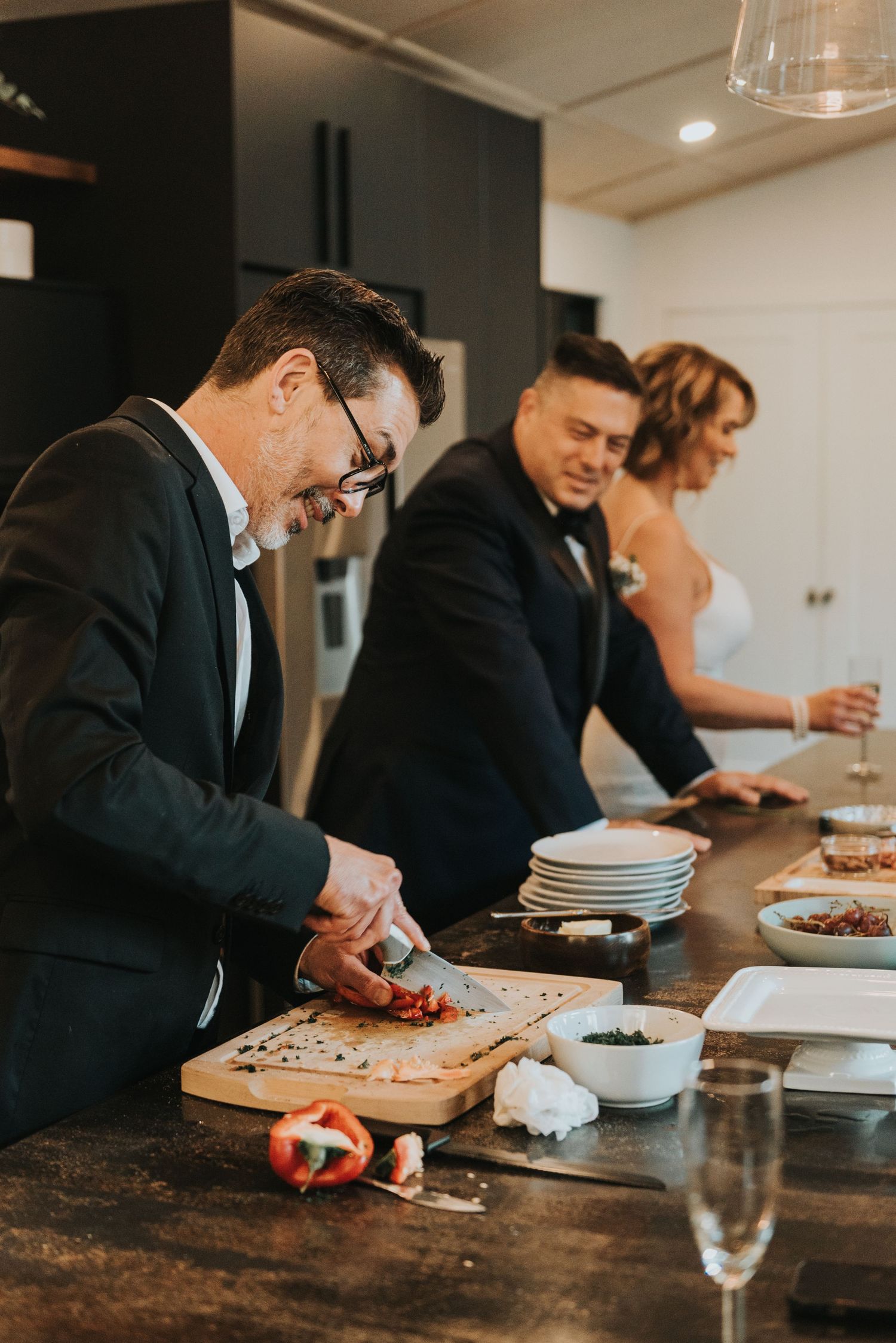 People in formal wear prepare food together at a wooden counter in a stylish kitchen setting.