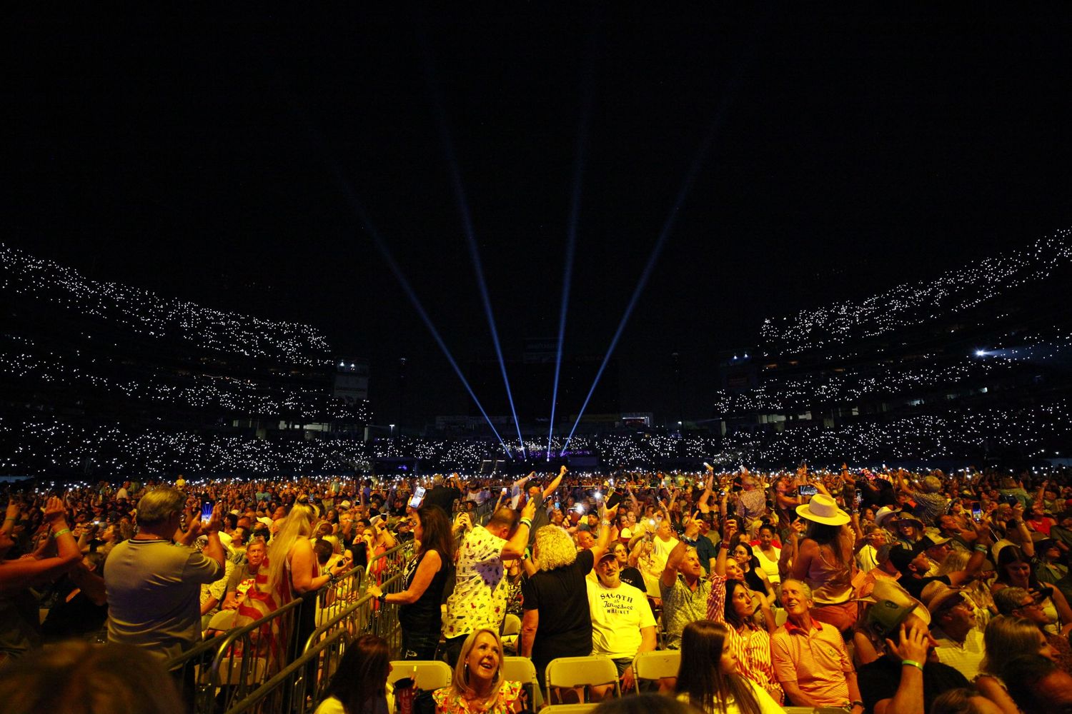 Large concert audience illuminated by yellow stage lights during nighttime outdoor performance.