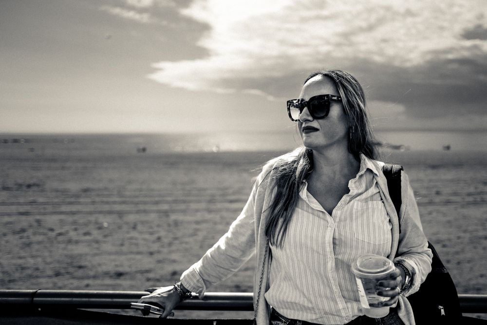 A black and white photo shows someone in sunglasses and striped shirt gazing at an ocean view during sunset.