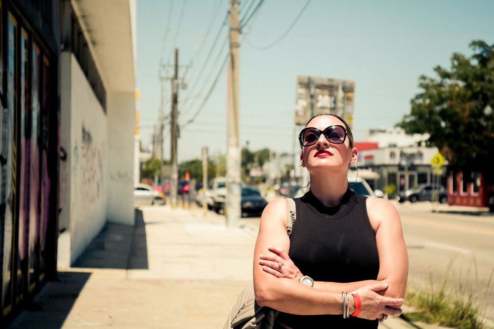 A person in sunglasses and black sleeveless top stands against a white wall on a sunny urban street.