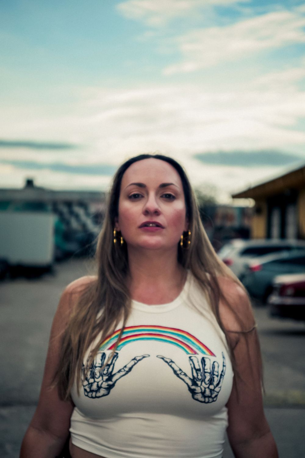 A white t-shirt features a colorful rainbow design between skeleton hand graphics in an urban parking lot setting.