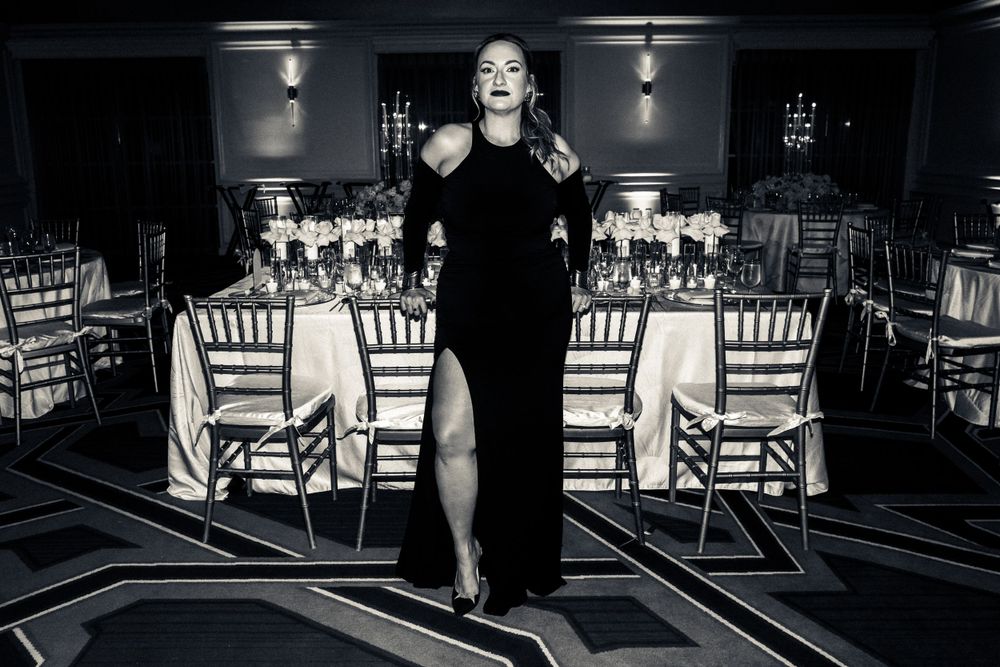 Black and white photo of an elegant figure in a long black evening gown standing in front of formal dining tables.