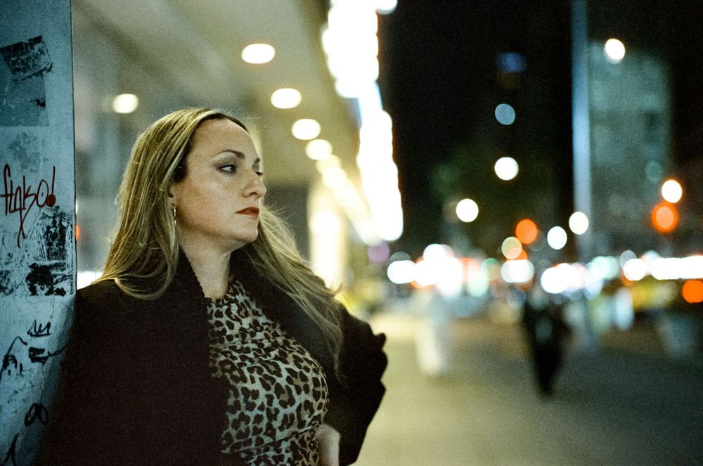 A person in a leopard print top and black coat stands on a city street at night with blurred lights in the background.