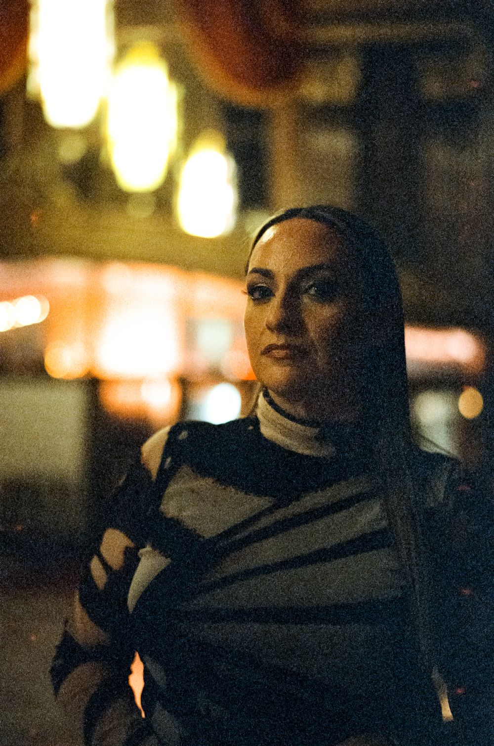 A woman in a dark sweater poses thoughtfully at night with blurred lights creating an atmospheric mood.