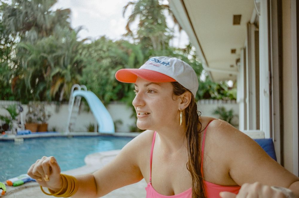 A person in a pink swimsuit and gray cap lounges poolside at a tropical resort with palm trees in the background.