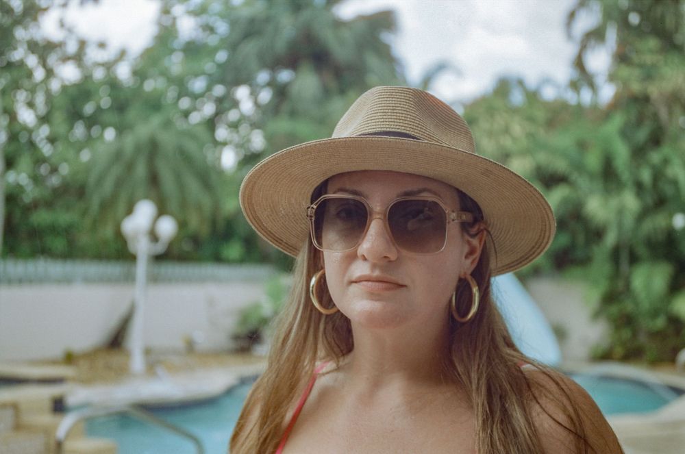 Woman wearing a straw sun hat and aviator sunglasses poses by a swimming pool on a summer day.