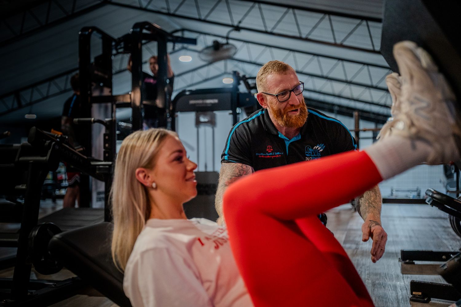 Personal trainer assists client during leg exercise workout at modern fitness gym facility.