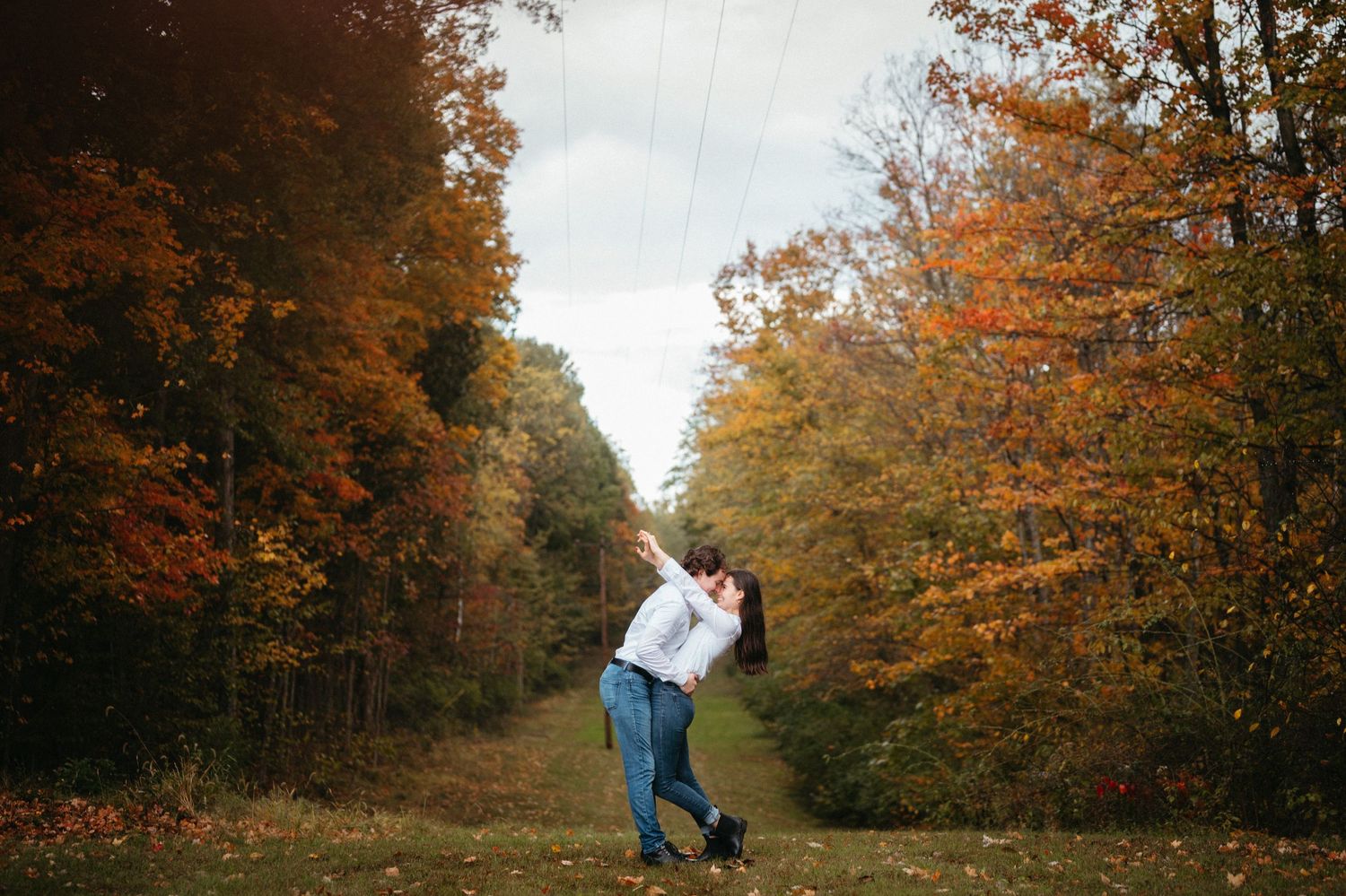 A couple dancing playfully on a path surrounded by vibrant autumn trees with orange and yellow foliage.
