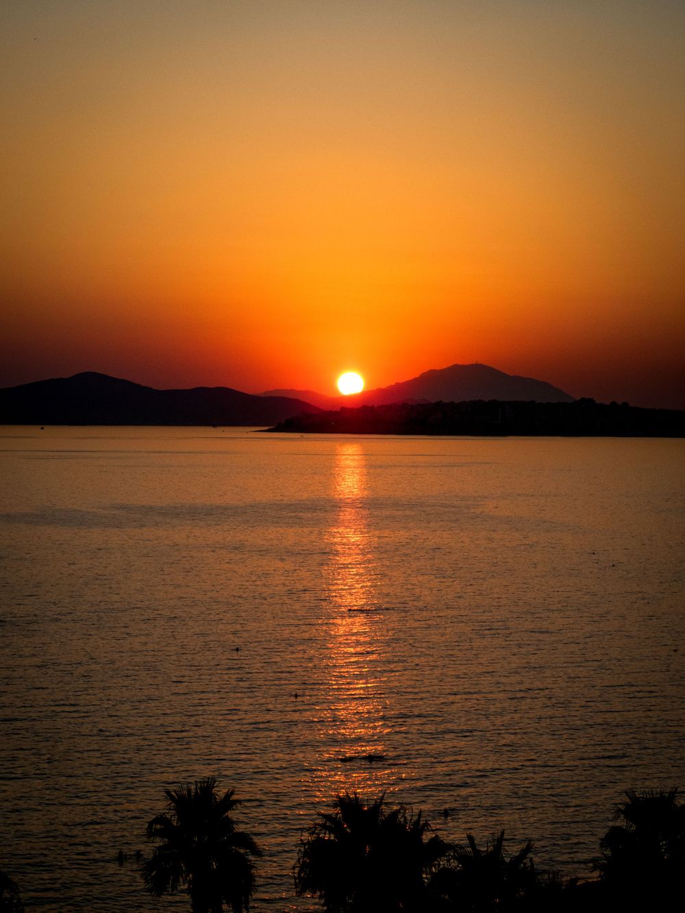Dramatic orange sunset over mountain silhouettes with reflection on calm water.