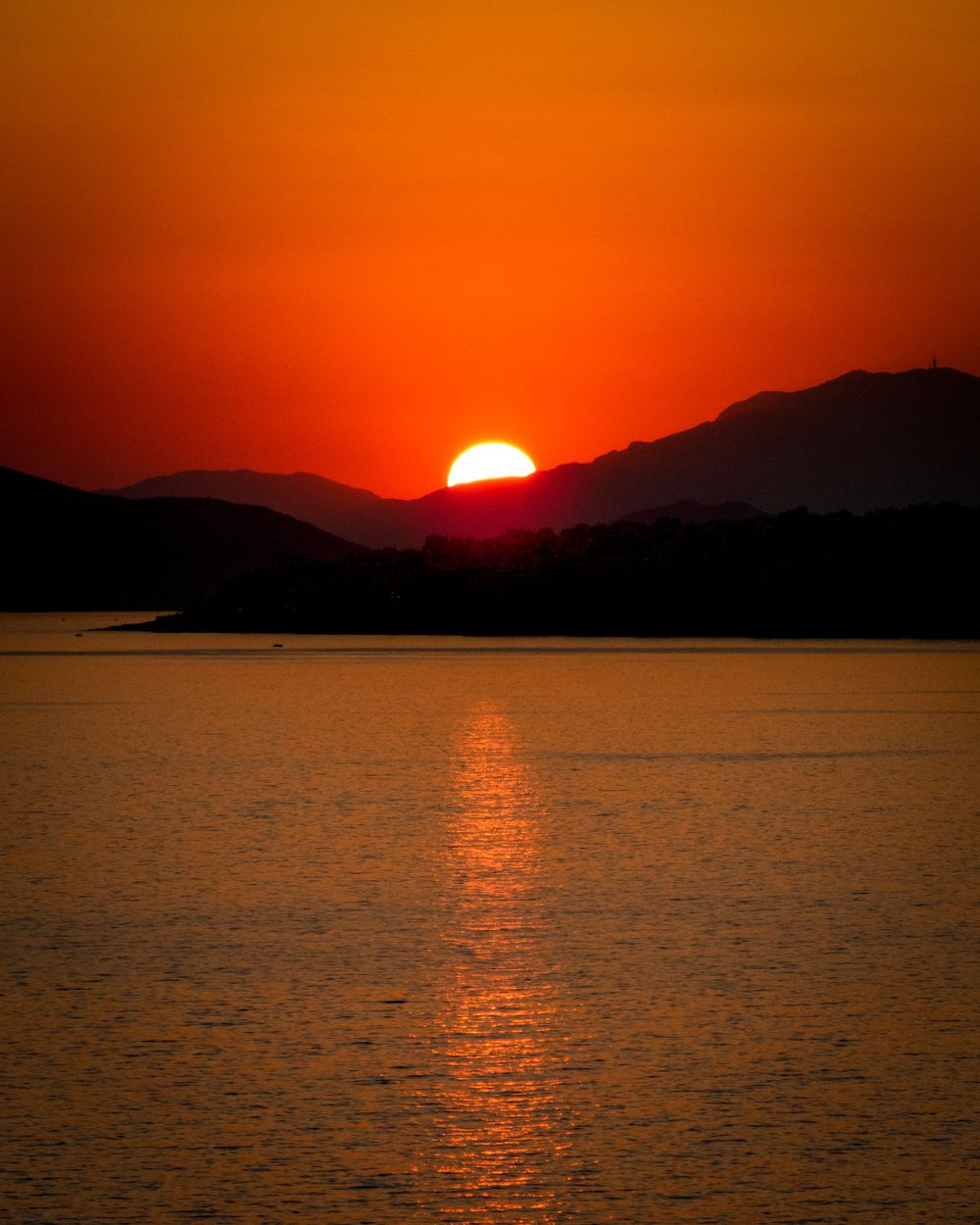 Vibrant red sunset sinking behind mountain range with golden reflection on water surface.