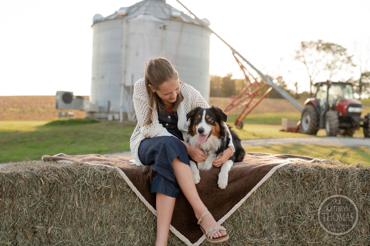 Person and dog share a moment on hay bale during golden hour with grain silo background.
