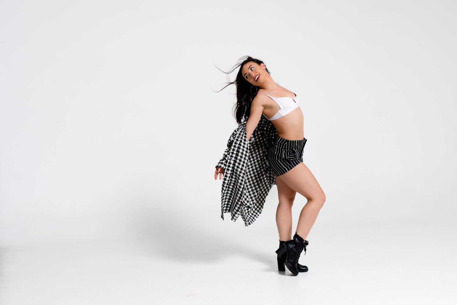 Dancer in black boots and shorts strikes dynamic pose against white background while wearing striped jacket.