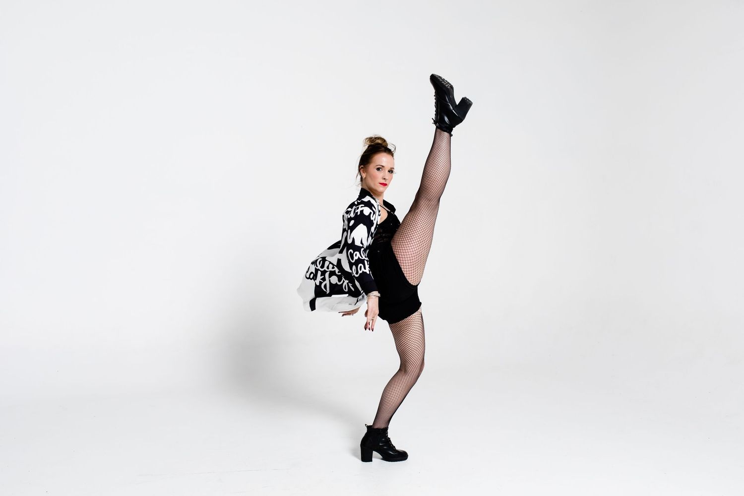 Dancer in black leotard and patterned jacket performs high kick against white background.