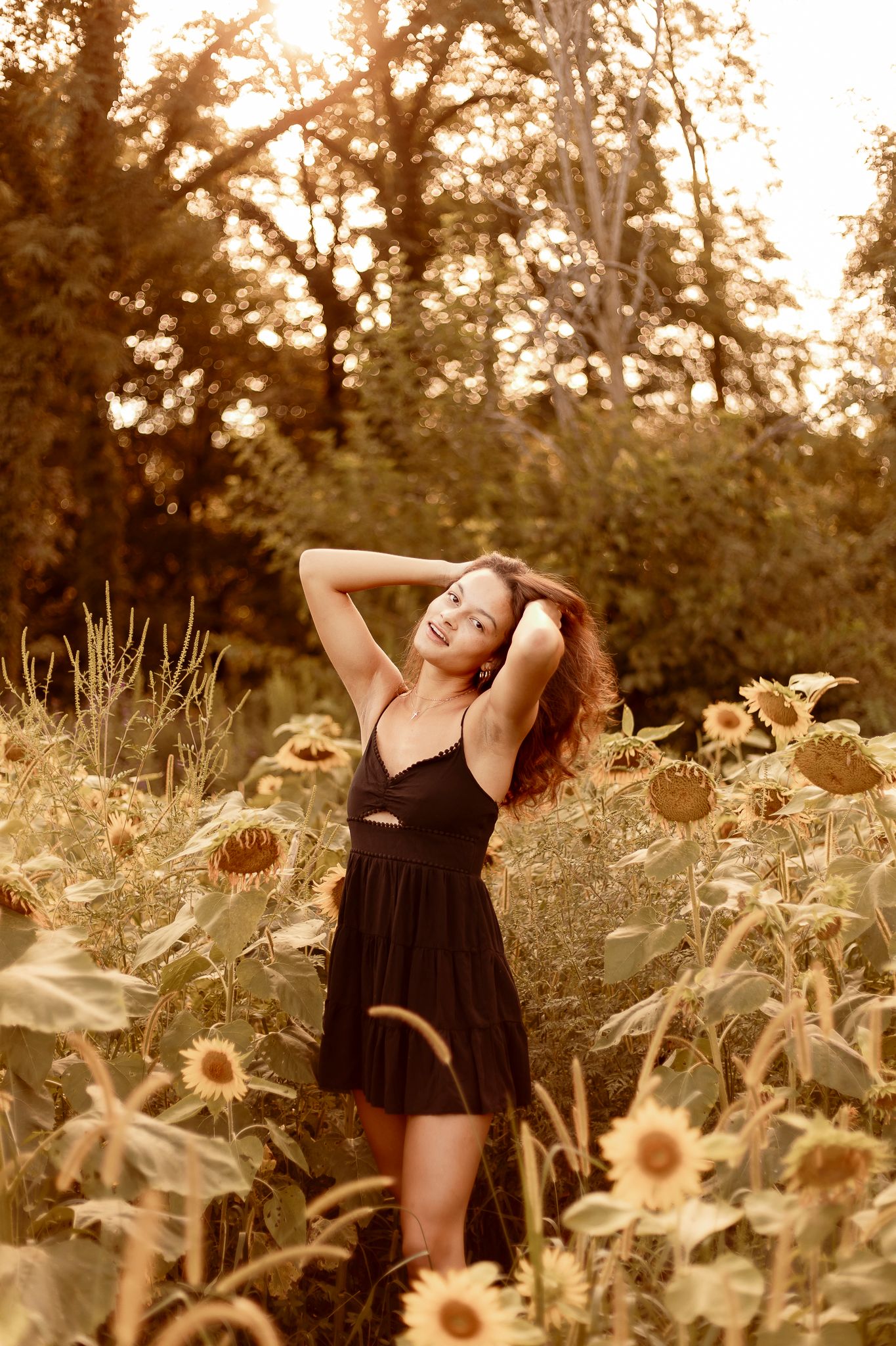 A woman in a black dress poses gracefully among blooming sunflowers at golden hour in a garden setting.