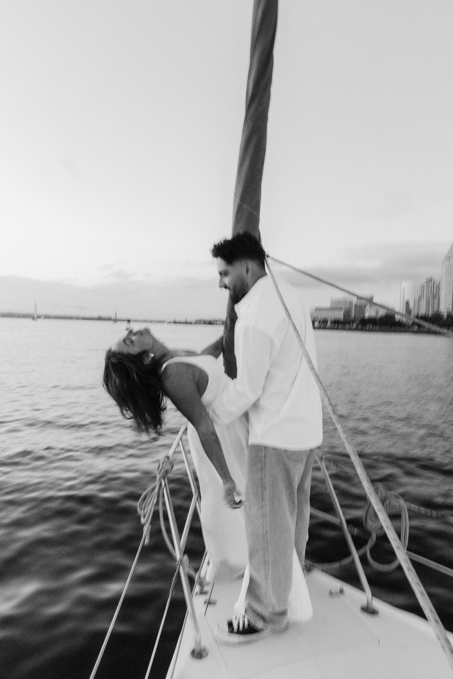 A romantic black and white scene of a couple embracing on the bow of a boat with a city skyline in the distance.