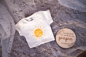 Baby onesie with yellow sun design next to wooden milestone marker on lace fabric.