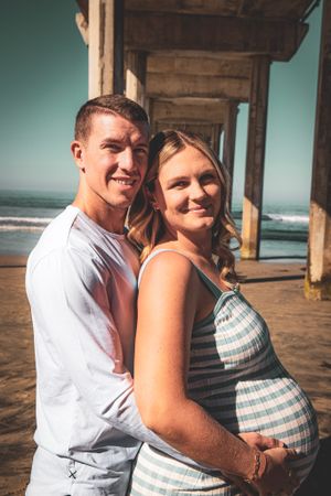 A couple poses together on a beach pier with the pregnant woman wearing a striped dress.