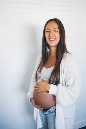 Joyful maternity portrait in white cardigan against light background.