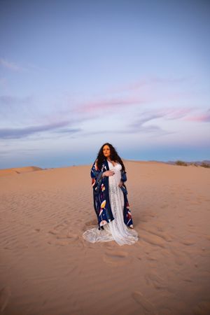 Elegant maternity portrait in a white gown and blue robe amid desert sand dunes.