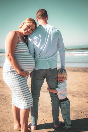A family shares a tender moment on the beach with their young child hugging legs.