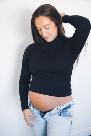 Side profile of expectant mother in black turtleneck and jeans against white wall.