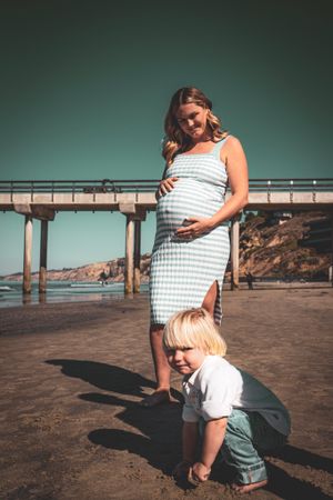 A pregnant woman in a striped dress stands by a pier while a child plays in the sand.