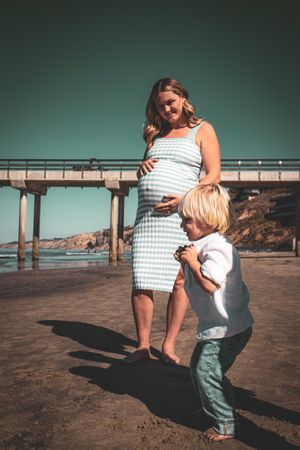A pregnant woman and child stand together near a beach pier on a sunny day.