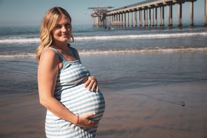 A pregnant woman in a striped dress smiles while standing on a beach with pier in background.