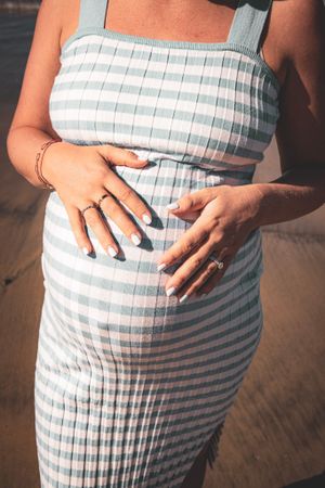 Close up of a pregnant belly in a blue and white striped maternity dress on the beach.
