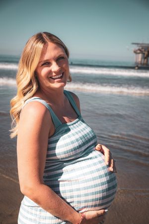 A pregnant woman poses joyfully on a beach wearing a striped dress.