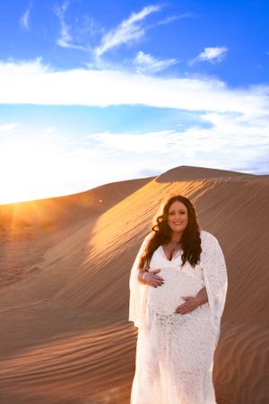Dreamy desert maternity photo with white lace dress against golden sand dunes at sunset.