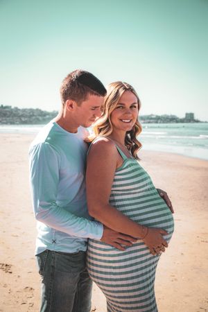 A couple embraces on a sunny beach while showing off her pregnant belly in a striped dress.