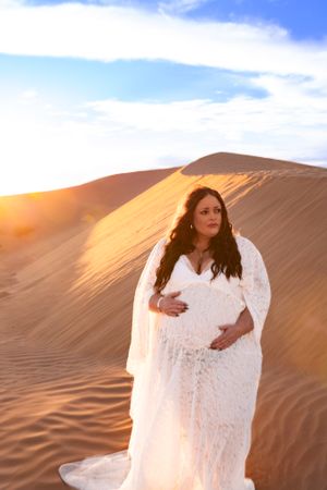 Maternity portrait in white lace against sweeping desert landscape during golden hour.