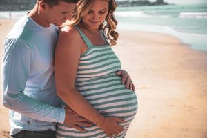 Expectant couple shares an intimate moment on a beach while wearing a striped maternity dress and casual attire.