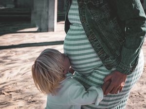 Child hugging pregnant mother wearing striped dress at beach.