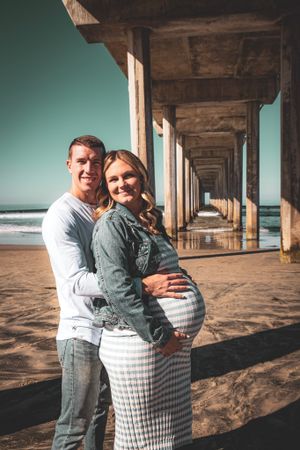 Maternity photo of couple embracing under beach pier at sunset.