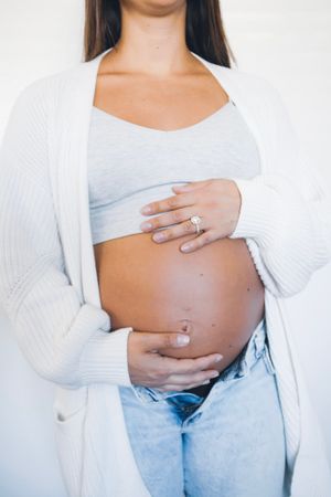 Pregnant woman in white cardigan and jeans showing baby bump with engagement ring.