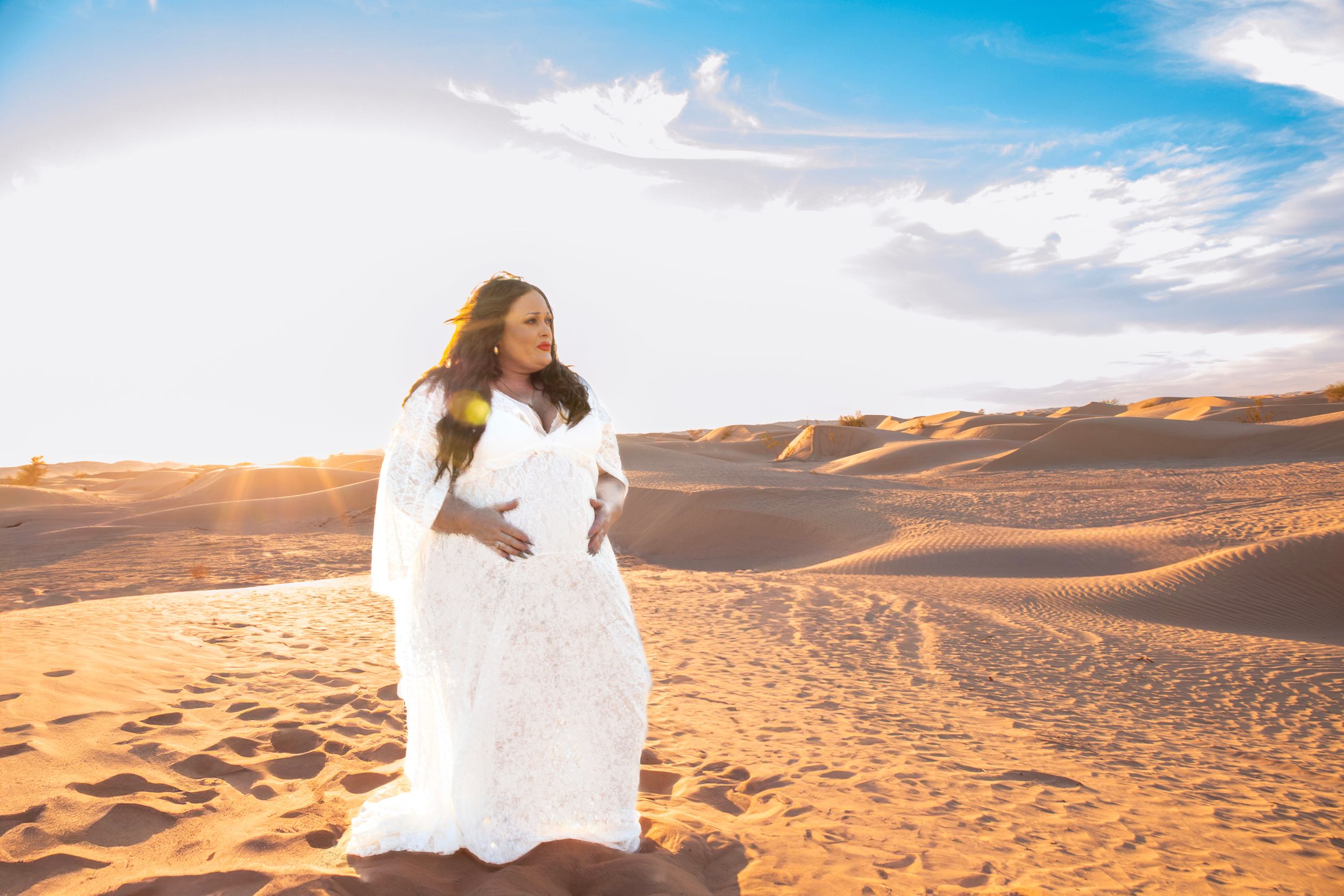 Desert maternity photography with billowing white dress against vast sand dunes.