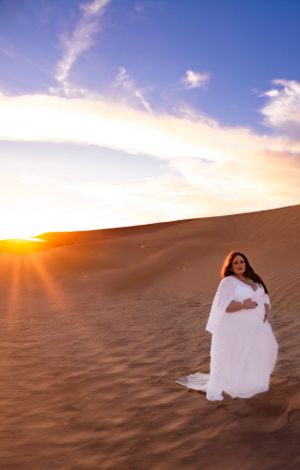 Dramatic sunset maternity photo with flowing white dress in desert sand dunes.