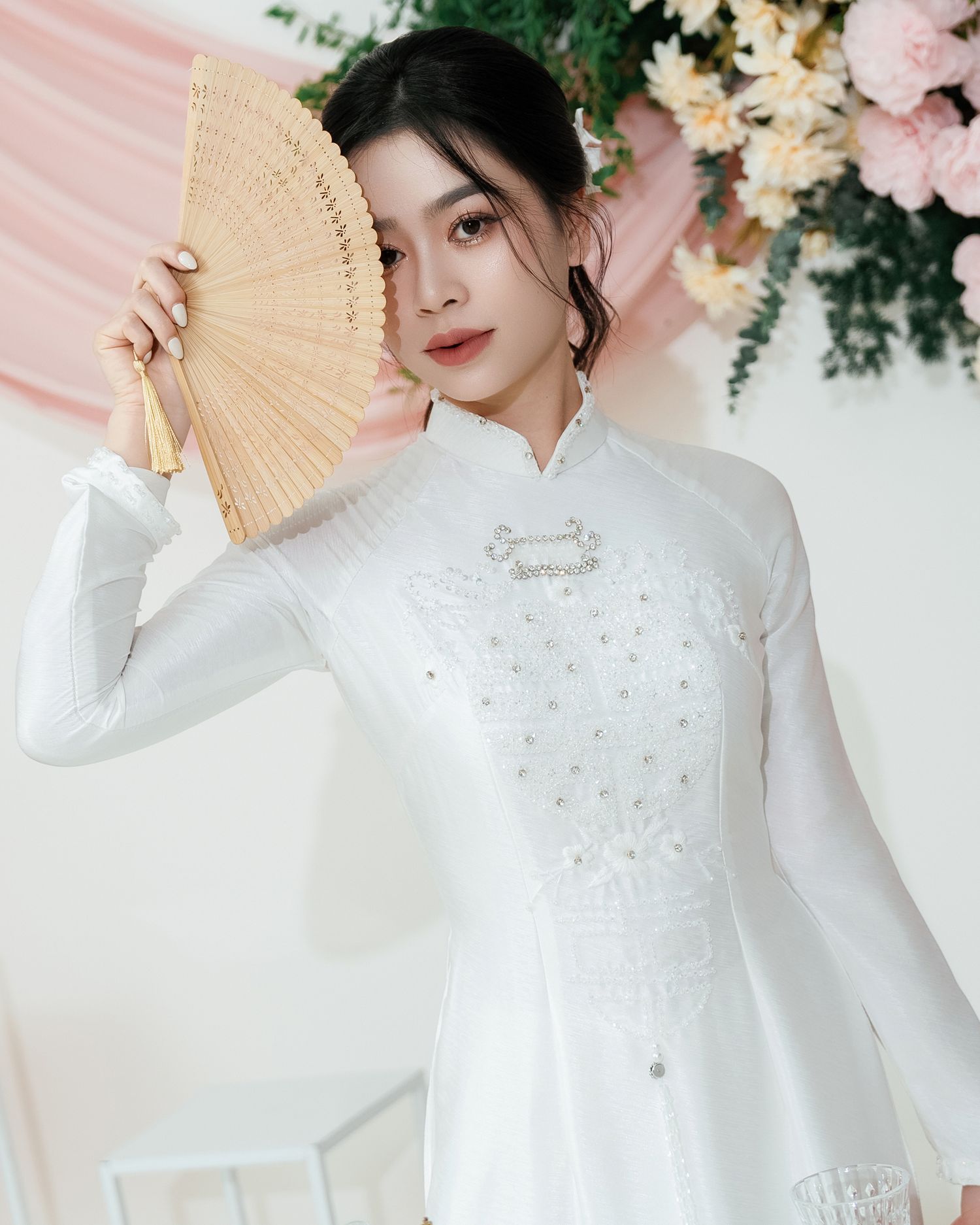 Traditional white Ao Dai dress with embellished details and a delicate handheld folding fan against floral setting.