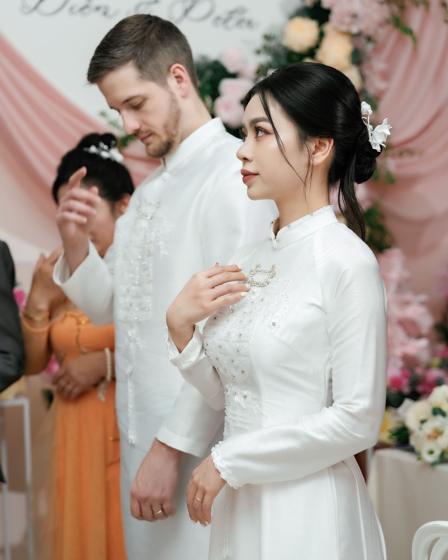 A traditional wedding ceremony with the couple in elegant white attire standing before pink floral decorations.