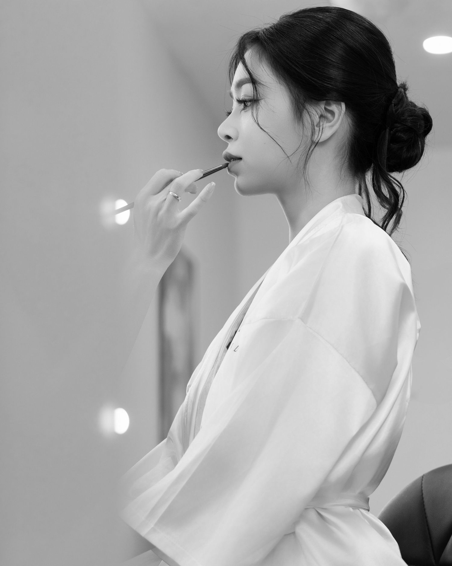 Black and white side profile photo showing someone in a white robe getting makeup applied during a beauty preparation.